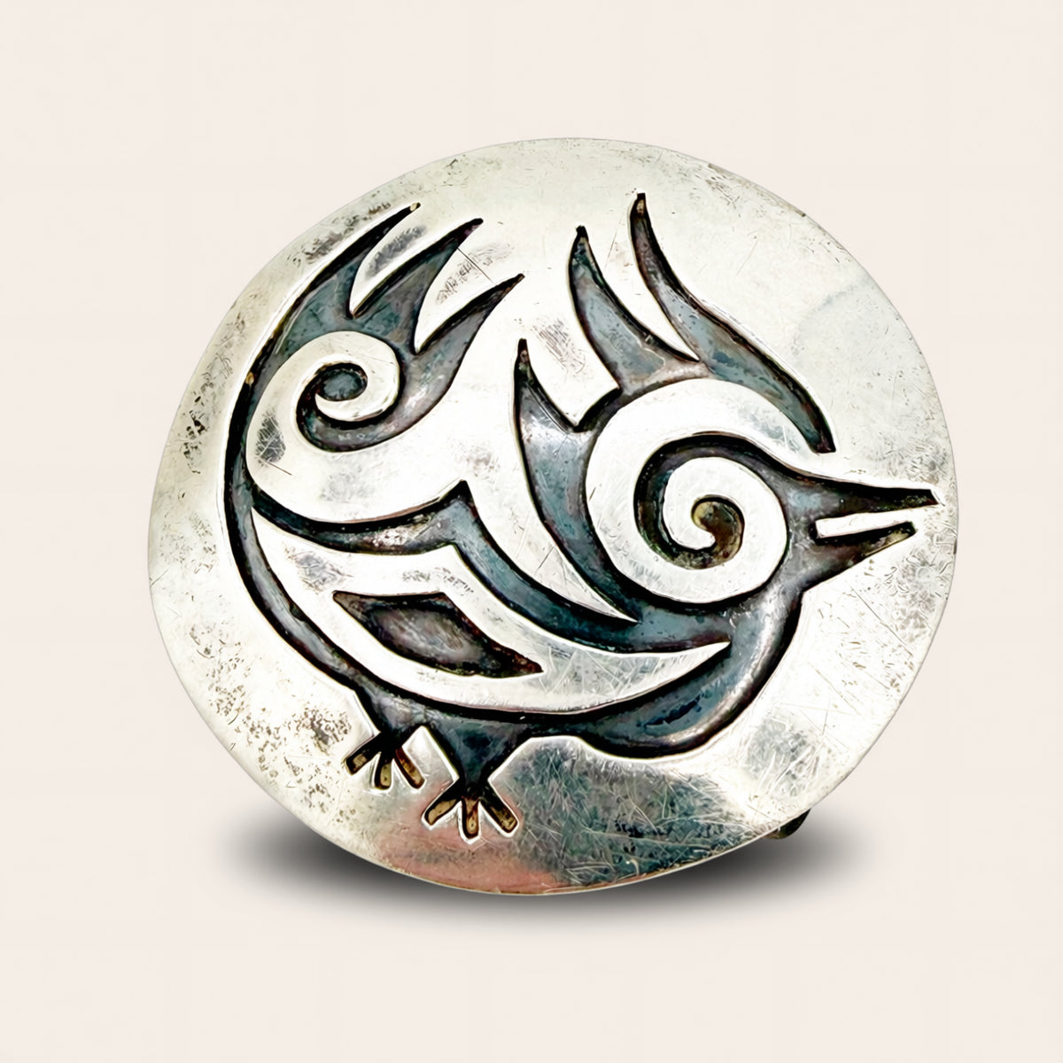 A round sterling silver belt buckle featuring a stylized bird design with swirling details and darkened accents, set against a light beige background.