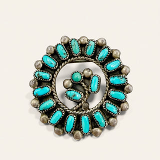 A circular silver-toned jewelry piece featuring multiple oval turquoise stones arranged in concentric rings, photographed on a plain light beige background.