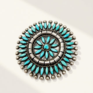 A circular silver-toned jewelry piece featuring turquoise stones arranged in concentric floral and geometric patterns, photographed on a clean off-white background with soft shadows.