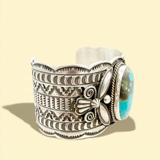 Silver cuff bracelet with intricate designs and a turquoise stone on a beige background