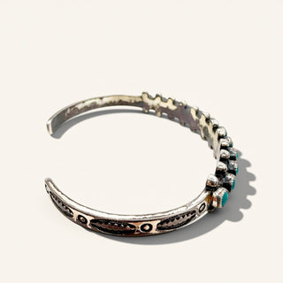 A sterling silver cuff bracelet with hand-hammered textures, engraved details, and a row of small turquoise beads on one end, shown on a light beige background.