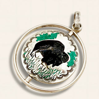 A circular sterling silver pendant featuring a black bison inlay, green grass and tree accents, and a textured silver border, shown on a light beige background.