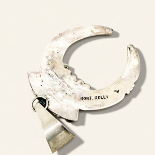 A silver crescent moon pendant with a triangular drop, featuring the engraved name 'ROBT. KELLY' on the main crescent, set against a light beige background.