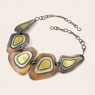 A statement necklace featuring five organic-shaped bronze-toned metal plates with gold and silver inlay, connected by a dark metal chain on a light beige background.