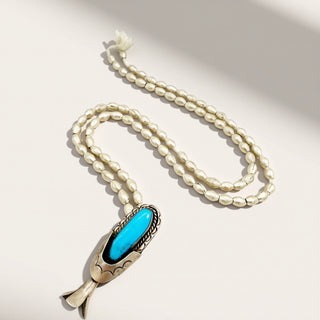 A silver-toned beaded necklace with a pendant featuring a bright turquoise blue stone, displayed on a light beige background with soft shadows.