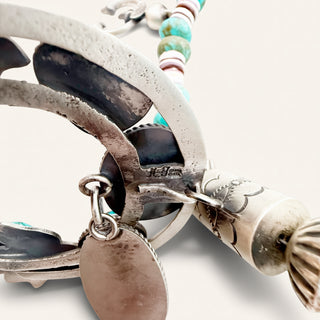 A close-up of a silver-toned Southwestern-style pendant necklace featuring turquoise and white bead details, a dangling oval tag, and engraved floral elements on a cylindrical bead.