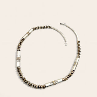 A silver-toned beaded necklace with textured cylindrical beads and polished accent tubes, arranged in a circle on a light beige background.