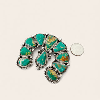 A silver-toned Southwestern-style turquoise pendant with multiple teardrop-shaped green stones and gold matrix details, displayed next to a US quarter coin for scale on a light beige background.
