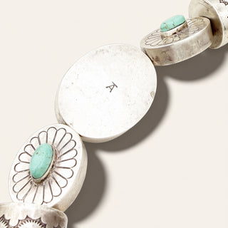 A close-up of a silver-toned jewelry piece with oval links and round floral-style end pieces set with turquoise stones, photographed on a plain light beige background.