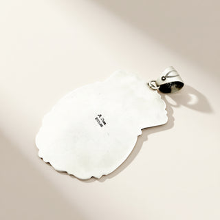 A sterling silver pendant with an organic, irregularly shaped body and a small bail at the top, shown on a warm beige background with soft shadows.