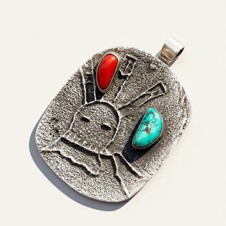 A textured sterling silver pendant featuring a hand-carved figure with two inlaid stones, one red and one turquoise, on a light cream background.