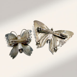 Two butterfly-shaped brooches in silver-tone metal, one showing the front and the other the back with pin clasp, on a light cream background.