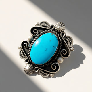 A silver-toned ornate ring featuring a large oval turquoise stone, photographed on a light surface with strong diagonal shadows.