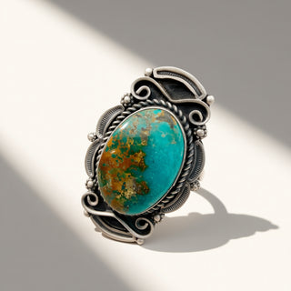 A silver-toned ring featuring a large oval turquoise stone with natural brown and gold matrix patterns, set in an ornate filigree-style band on a light gray background with soft diagonal shadows.