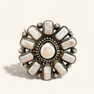 An ornate circular silver-toned cuff featuring a central irregular white pearl surrounded by multiple rectangular white mother-of-pearl stones, set against a light beige background.
