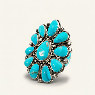 A large silver-toned cuff featuring multiple turquoise stones arranged in a floral cluster, photographed on a clean off-white background.
