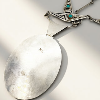 A close-up of a silver pendant necklace featuring a large oval silver disc pendant and a geometric silver chain with turquoise and silver bead details, set against a plain light background.