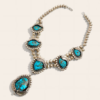 A silver-toned Southwestern-style necklace featuring five turquoise-colored stones in ornate bezel settings, displayed on a light beige background.