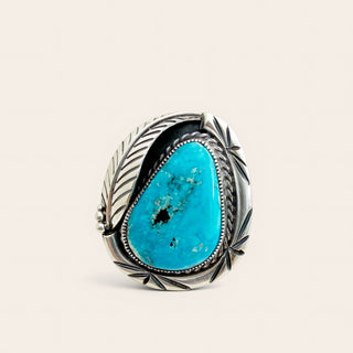 A sterling silver ring featuring a large, teardrop-shaped turquoise stone with natural matrix inclusions, set in an ornate, engraved bezel with leaf-like details on a light cream background.