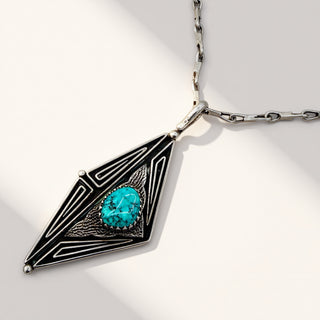 A silver pendant necklace with a geometric diamond-shaped design, featuring black enamel inlay and a central oval turquoise gemstone, shown on a light gray background.