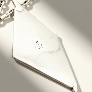 A close-up of a silver pendant with a geometric triangular shape, featuring a small engraved symbol, shown on a light background with dramatic shadows.
