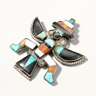 A silver-toned Native American style inlay brooch featuring geometric turquoise, black, white, and orange stone sections, photographed on a light cream background.