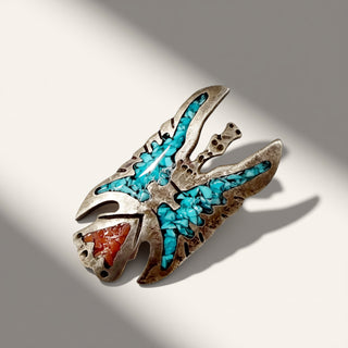 A silver-toned Native American style pendant featuring turquoise inlay and a red-orange stone accent, photographed on a light gray background with dramatic diagonal shadows.