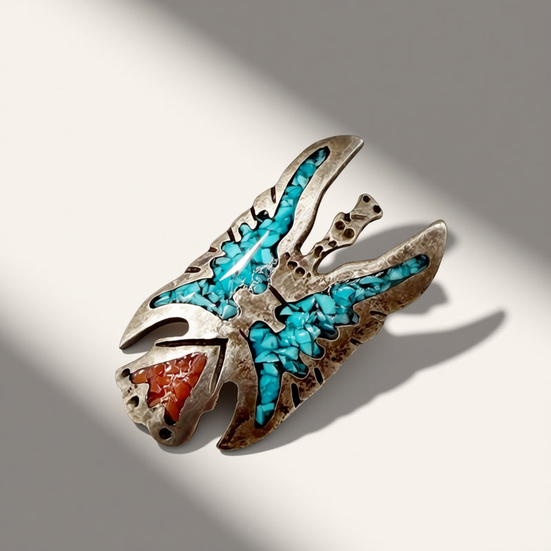 A silver-toned Native American style pendant featuring turquoise inlay and a red-orange stone accent, photographed on a light gray background with dramatic diagonal shadows.