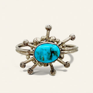 A sterling silver cuff ring featuring a central oval turquoise cabochon with organic dark inclusions, surrounded by textured, branching silver spikes on a light cream background.