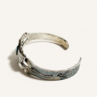 A sterling silver cuff bracelet with hand-stamped geometric and bird motifs, featuring a small turquoise stone accent on the left side, photographed on a clean off-white background.