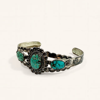 An ornate silver cuff bracelet featuring three oval turquoise stones, set against a plain off-white background.