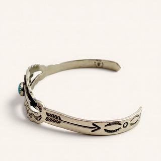A sterling silver cuff bracelet with hand-stamped Navajo-style arrow and geometric designs, featuring a small turquoise stone accent on the left side, photographed on a clean off-white background.