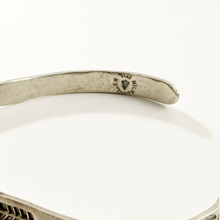 Close-up of a silver-toned metal cuff bracelet showing engraved details and a small heart-shaped mark on the inner band, photographed on a clean white background.