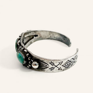 A silver-toned open cuff bracelet with engraved geometric and arrow motifs, accented by a round green stone and a small metallic bead on the front, photographed on a light cream background.
