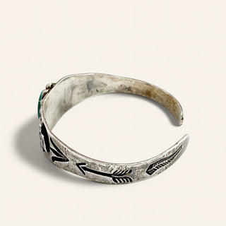 A silver-toned open cuff bracelet with engraved arrow motifs and a small green stone accent on the left side, photographed on a plain light beige background.