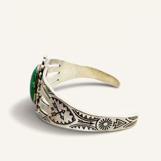 A sterling silver cuff ring with intricate hand-stamped geometric and floral patterns, featuring a round green cabochon stone on the side, set against a plain off-white background.