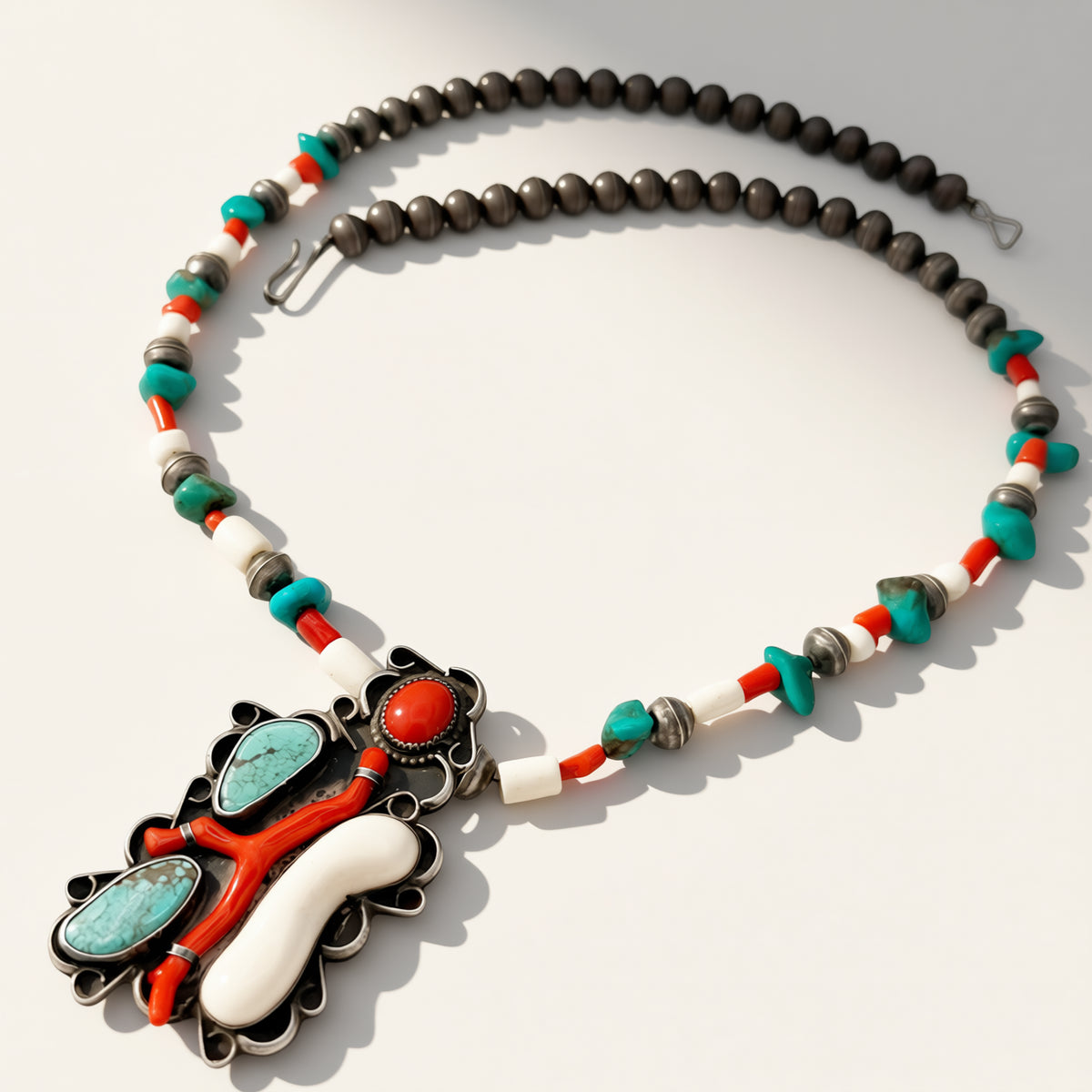 A Southwestern-style beaded necklace with a large pendant, featuring turquoise, white, and red beads alongside dark gray beads, on a light gray background.