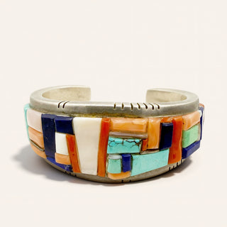 A Southwestern-style cuff bracelet featuring a silver-tone metal band decorated with a geometric mosaic of turquoise, coral, white, and dark blue stone inlays, set against a plain off-white background.