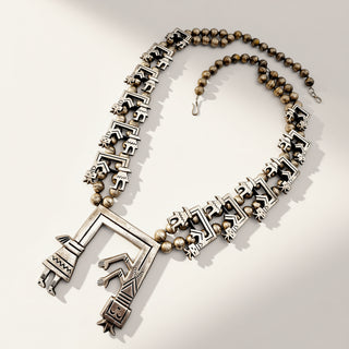 A silver-toned statement necklace featuring a large geometric pendant with stylized human and architectural motifs, flanked by rows of small animal-shaped charms and accented with round beads, displayed on a light beige background.