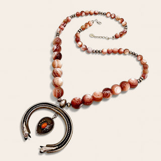 A beaded necklace featuring round agate-like beads in shades of terracotta, rust, cream, and brown, accented with small metallic spacer beads. The necklace hangs from a large silver-toned circular pendant with a small teardrop orange stone, photographed on a plain light beige background.