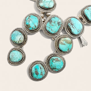 A close-up of a silver-toned jewelry piece featuring multiple oval turquoise stones set in ornate braided silver bezels, photographed on a plain light beige background.