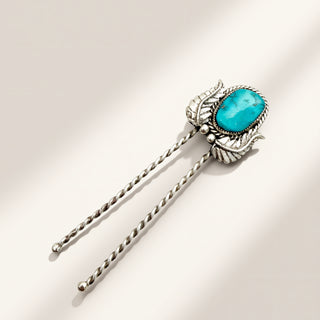 A sterling silver hair stick with a decorative leaf-shaped setting holding a vibrant turquoise stone, shown on a warm beige background with soft diagonal shadows.