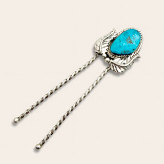 A sterling silver sash pin with a vibrant turquoise stone in a leaf-shaped setting, featuring two twisted metal prongs, shown on a light beige background.
