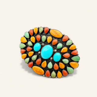A Southwestern-style oval turquoise and coral brooch with a black metal setting, featuring a large central turquoise cabochon, two smaller turquoise stones, and multiple coral and turquoise stones in orange, green, and red-orange tones.