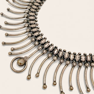 An ornate antique-style silver-toned collar necklace with dangling curved bars, beadwork, and a large ring pendant, shown on a plain light beige background.