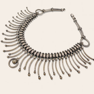 An antique-style silver  collar necklace with dangling curved bars, and a small round pearl accent, displayed on a light beige background.