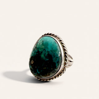 A silver ring featuring a large, organic-shaped turquoise cabochon stone with natural dark inclusions, set in a braided silver bezel, photographed on a light cream background.