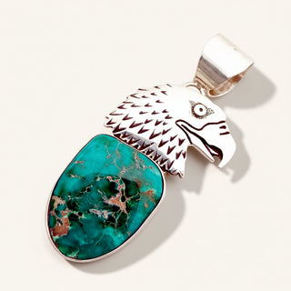 A sterling silver eagle head pendant featuring a large, polished turquoise stone with natural brown matrix veining, set against a clean, light beige background.