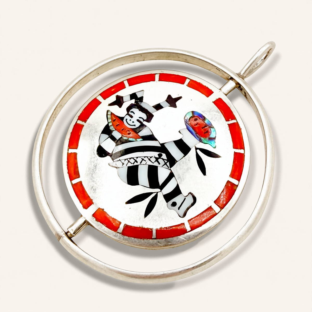 A circular silver-toned pendant featuring a hand-painted Inuit-style design with a black-and-white striped figure, a red border, and small colorful details, shown on a light beige background.