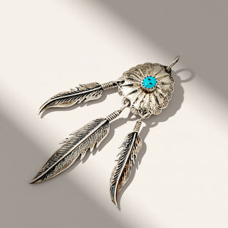 A sterling silver dreamcatcher pendant with three hanging feathers and a central turquoise stone, photographed on a light beige background with soft shadows.
